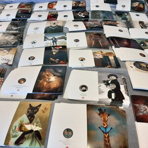 Puede incluir: Una gran pila de tarjetas de felicitaci&oacute;n blancas con varias ilustraciones de animales. Las tarjetas presentan una variedad de animales, incluidos gatos, perros, una jirafa y un ping&uuml;ino. Las tarjetas son todas blancas con un logotipo de hoja de arce roja en la esquina inferior derecha.