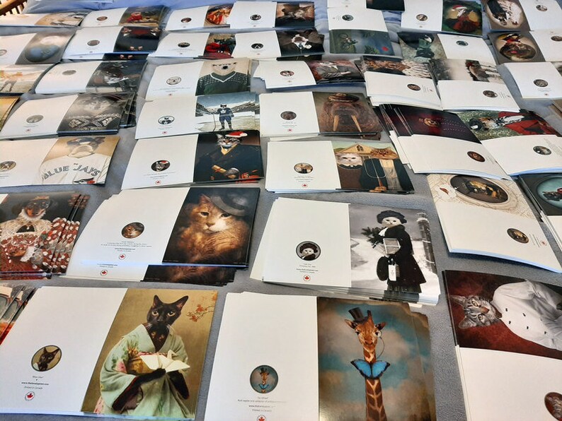 Puede incluir: Una gran pila de tarjetas de felicitaci&oacute;n blancas con varias ilustraciones de animales. Las tarjetas presentan una variedad de animales, incluidos gatos, perros, una jirafa y un ping&uuml;ino. Las tarjetas son todas blancas con un logotipo de hoja de arce roja en la esquina inferior derecha.