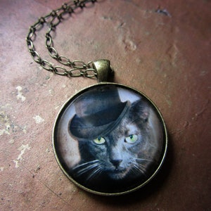 May include: A silver chain necklace with a round pendant featuring a black cat wearing a top hat. The cat has green eyes and a white mustache.