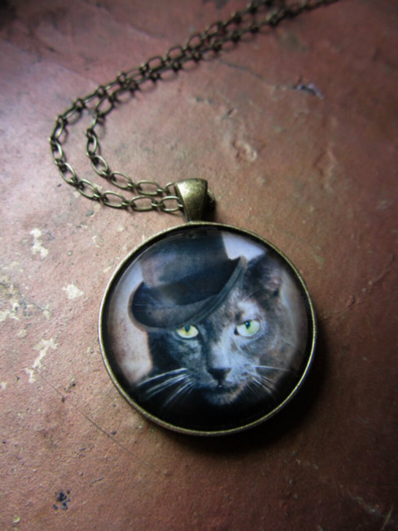 May include: A silver chain necklace with a round pendant featuring a black cat wearing a top hat. The cat has green eyes and a white mustache.