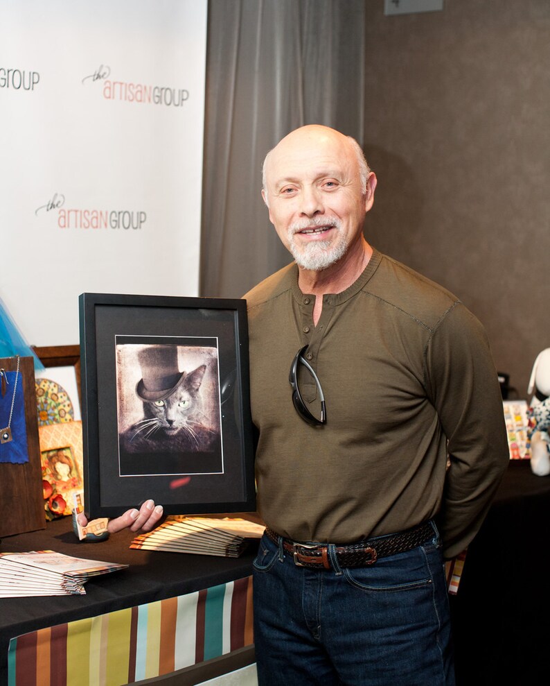 May include: A man in a brown long-sleeved shirt and blue jeans is holding a framed black and white photograph of a cat wearing a top hat. The photograph is on a table with a stack of brochures and a blue necklace.