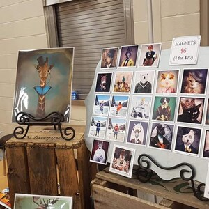 Peut inclure: Un pr&eacute;sentoir de tirages encadr&eacute;s et d'aimants illustrant des animaux. Les tirages sont sur des chevalets en m&eacute;tal noir et les aimants sont sur un tableau blanc. Les aimants co&ucirc;tent 6 &euro; pi&egrave;ce ou 4 pour 20 &euro;. Les tirages co&ucirc;tent 10 &euro; pi&egrave;ce ou 3 pour 25 &euro;. Les animaux comprennent une girafe, un chat, un &eacute;lan, un chien et un ours.