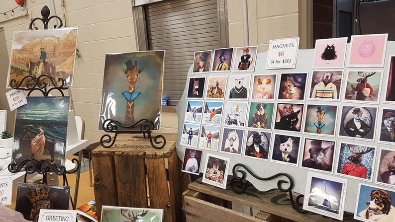 Peut inclure: Un pr&eacute;sentoir de tirages encadr&eacute;s et d'aimants illustrant des animaux. Les tirages sont sur des chevalets en m&eacute;tal noir et les aimants sont sur un tableau blanc. Les aimants co&ucirc;tent 6 &euro; pi&egrave;ce ou 4 pour 20 &euro;. Les tirages co&ucirc;tent 10 &euro; pi&egrave;ce ou 3 pour 25 &euro;. Les animaux comprennent une girafe, un chat, un &eacute;lan, un chien et un ours.