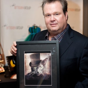 May include: A man in a suit holds a framed black and white photograph of a cat wearing a top hat. The cat has green eyes and a serious expression.