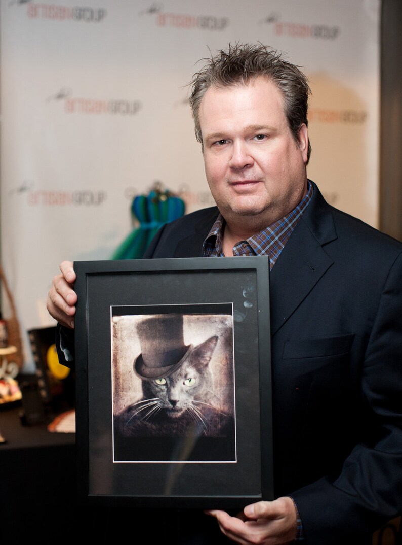 May include: A man in a suit holds a framed black and white photograph of a cat wearing a top hat. The cat has green eyes and a serious expression.