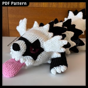Puede incluir: Un juguete de dragón de ganchillo hecho a mano en blanco, negro y rosa. El dragón tiene una cabeza blanca con detalles negros, una lengua rosa y un cuerpo negro y blanco. El texto "PDF Pattern" es visible en la esquina superior izquierda.