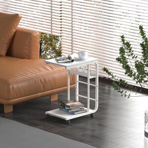 Puede incluir: Mesa auxiliar blanca con un dise&ntilde;o curvo y dos estantes. La mesa tiene ruedas y est&aacute; al lado de un sof&aacute; marr&oacute;n.