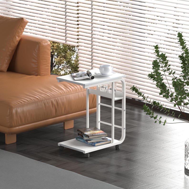 Puede incluir: Mesa auxiliar blanca con un dise&ntilde;o curvo y dos estantes. La mesa tiene ruedas y est&aacute; al lado de un sof&aacute; marr&oacute;n.