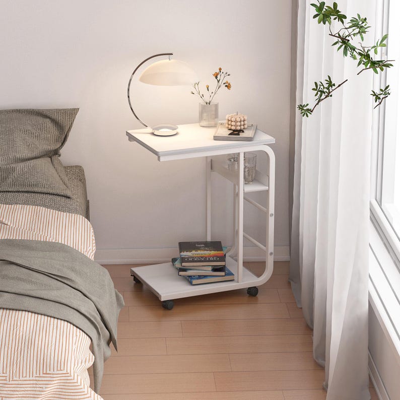 Puede incluir: Mesita de noche blanca con un estante y ruedas. La mesa tiene una l&aacute;mpara, un jarr&oacute;n con flores y un libro en la parte superior.