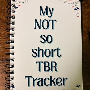 May include: A white spiral-bound notebook with the text "My NOT SO short TBR Tracker" in navy blue. The notebook is decorated with small book illustrations in red, blue, and green.