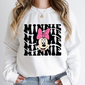 K&ouml;nnte beinhalten: Wei&szlig;er Pullover mit einem schwarz-wei&szlig;en Grafikdruck von Minnie Mauss Gesicht und dem Wort "Minnie", das in einem wellenf&ouml;rmigen Muster wiederholt wird.