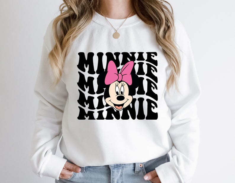 K&ouml;nnte beinhalten: Wei&szlig;er Pullover mit einem schwarz-wei&szlig;en Grafikdruck von Minnie Mauss Gesicht und dem Wort "Minnie", das in einem wellenf&ouml;rmigen Muster wiederholt wird.
