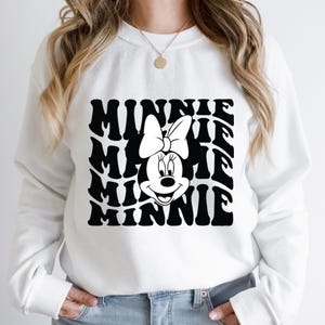 K&ouml;nnte beinhalten: Wei&szlig;er Pullover mit einem schwarz-wei&szlig;en Motiv von Minnie Maus mit dem Wort "Minnie", das in einer wellenf&ouml;rmigen Form wiederholt wird.