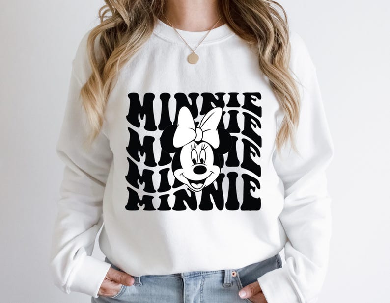 K&ouml;nnte beinhalten: Wei&szlig;er Pullover mit einem schwarz-wei&szlig;en Motiv von Minnie Maus mit dem Wort "Minnie", das in einer wellenf&ouml;rmigen Form wiederholt wird.