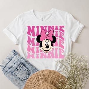 K&ouml;nnte beinhalten: Wei&szlig;es T-Shirt mit einer pinkfarbenen und wei&szlig;en Grafik von Minnie Maus mit dem Wort "Minnie", das in einer Retro-Schrift wiederholt wird.