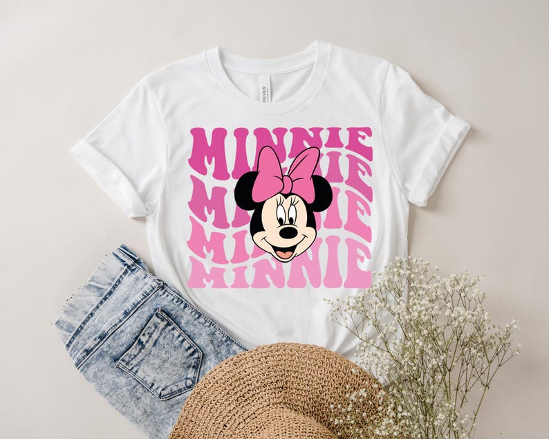 K&ouml;nnte beinhalten: Wei&szlig;es T-Shirt mit einer pinkfarbenen und wei&szlig;en Grafik von Minnie Maus mit dem Wort "Minnie", das in einer Retro-Schrift wiederholt wird.