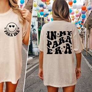 May include: A beige t-shirt with a black smiley face with hearts for eyes and the text "Para Era" on the front. The back of the shirt has the text "In My Para Era" in a retro font.