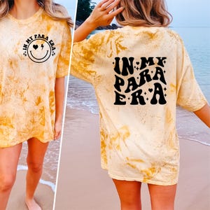 May include: A yellow tie-dye t-shirt with the text "In My Para Era" in black block letters with a heart and star design. The front of the shirt has a smiley face with a heart and lightning bolt.