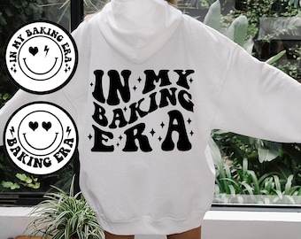 May include: White hoodie with a black graphic that says "In My Baking Era" with a smiley face with hearts for eyes and a lightning bolt above it.