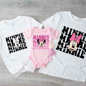 K&ouml;nnte beinhalten: Drei wei&szlig;e T-Shirts mit einer schwarz-wei&szlig;en Grafik von Minnie Maus mit dem Wort "Minnie", das in einem Retro-Stil wiederholt wird. Das kleinste Shirt ist rosa und hat kurze &Auml;rmel. Die beiden gr&ouml;&szlig;eren Shirts haben lange &Auml;rmel.