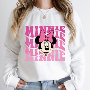K&ouml;nnte beinhalten: Wei&szlig;er Pullover mit einem pinkfarbenen und wei&szlig;en Aufdruck von Minnie Mauss Gesicht und dem Wort "Minnie", das in einer Retro-Schrift wiederholt wird.