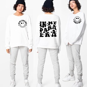 May include: White sweatshirt with long sleeves and a black graphic on the front that says "IN MY PARA ERA" in a retro font with hearts and stars. The back of the sweatshirt has a smiley face with hearts for eyes and the text "IN MY PARA ERA" in a smaller font.