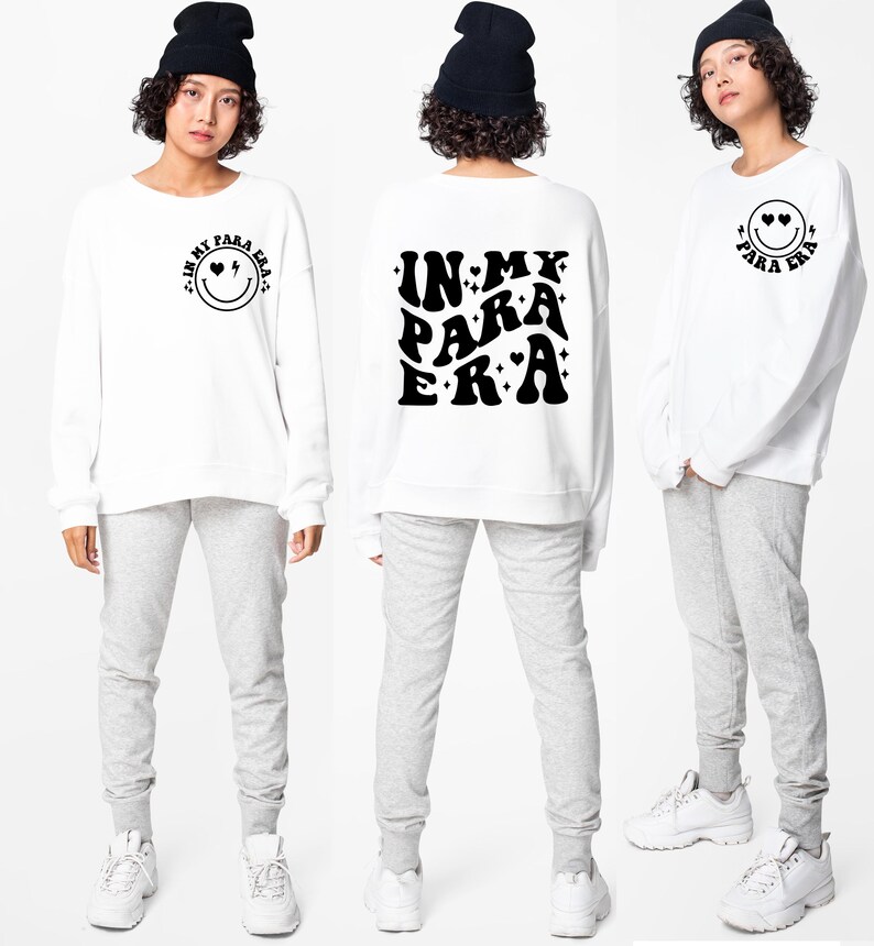 May include: White sweatshirt with long sleeves and a black graphic on the front that says "IN MY PARA ERA" in a retro font with hearts and stars. The back of the sweatshirt has a smiley face with hearts for eyes and the text "IN MY PARA ERA" in a smaller font.