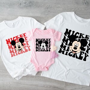 May include: Three white t-shirts with a black and white Mickey Mouse graphic and the word "Mickey" repeated in red and black. The shirts are in different sizes, with the smallest shirt being pink.