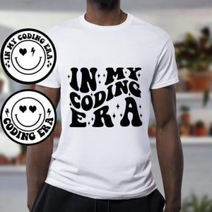 May include: White t-shirt with black text that reads "In My Coding Era" in a retro font. The text is surrounded by small stars. The shirt also features a black and white smiley face with hearts for eyes and a lightning bolt for a mouth.