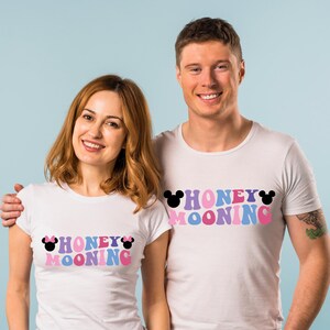May include: A couple wearing white t-shirts with the text "Honeymooning" in colourful letters and Mickey Mouse ears. The woman's shirt has pink Mickey Mouse ears and the man's shirt has black Mickey Mouse ears.
