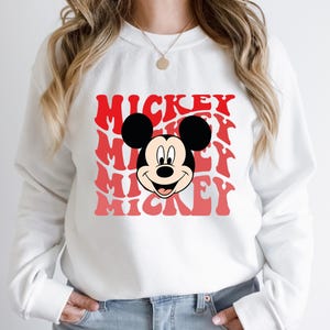 May include: White sweatshirt with a red and pink graphic of Mickey Mouse's face and the word "Mickey" repeated multiple times.