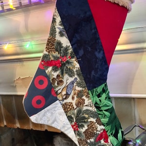 May include: A Christmas stocking made of patchwork fabric with a variety of holiday-themed prints, including pine cones, birds, and holly. The stocking is red, green, blue, and white.
