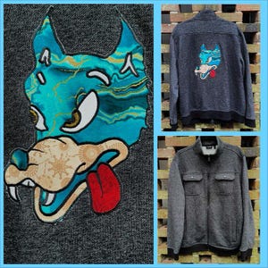 May include: A gray fleece jacket with a blue and gold wolf design on the back. The wolf has a red tongue sticking out. The jacket has a zipper closure and two pockets on the front.