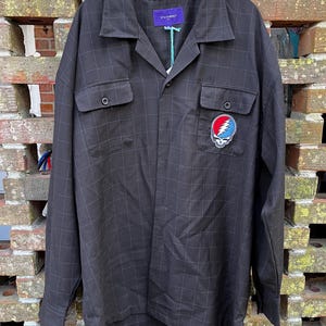 May include: Black button-up shirt with a subtle plaid pattern. Features two chest pockets and a colorful Steal Your Face patch on the right side. The shirt has a collar and is hanging on a black hanger. The tag reads "STATEMENT".