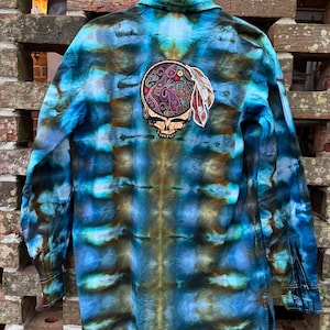May include: A long-sleeved button-down shirt with a tie-dye pattern in shades of blue, brown, and green. The back features a patch with a skull design, paisley details, and a feather. The shirt is hanging against a brick wall.