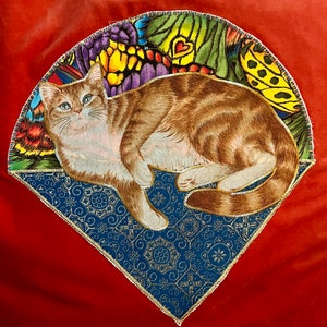 Puede incluir: Una almohada de tela roja con un diseño de gato bordado de colores. El gato está acostado con la cabeza girada hacia un lado. El bordado está rodeado de una tela estampada azul y dorada.