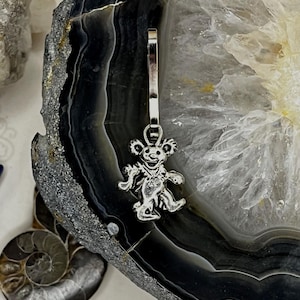 May include: A silver-colored charm in the shape of a dancing bear, attached to a silver clasp. The charm is detailed with facial features and a smiling expression. The background is a polished, multi-toned stone.