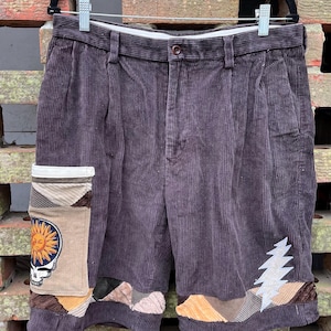 Può includere: Pantaloncini in velluto a coste viola scuro con chiusura a bottone e pieghe. I pantaloncini presentano dettagli patchwork, tra cui una grafica di sole e teschio e un fulmine. I pantaloncini sono appesi a una gruccia nera.