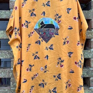 Könnte beinhalten: Senfgelbes Kimono-Top mit floralem Muster in Blau, Rot und Weiß. Ein zentraler Aufnäher zeigt eine schwarze Katzen-Silhouette auf blauem Hintergrund, eingebettet in ein Paisley-Design.