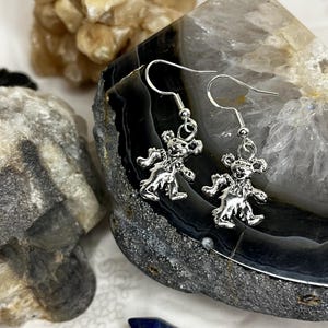 May include: Silver-toned bear earrings with a dancing bear design. The earrings are displayed on a dark gray and white stone surface, with a blue crystal and other stones in the background. The earrings have a hook closure.