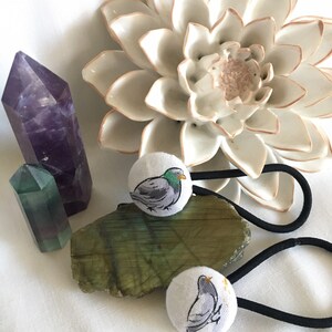 May include: Two white fabric hair ties with a gray pigeon illustration, set on a green stone with a purple crystal and a white ceramic flower. The hair ties have black elastic bands. The pigeon illustration has green and yellow accents.