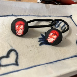 Puede incluir: Dos gomas para el pelo negras con botones circulares de tela negra que muestran un retrato de una persona con pelo rojo y un rayo en la cara. Los botones están sobre un plato de cerámica blanco con borde azul y un diseño de corazón.