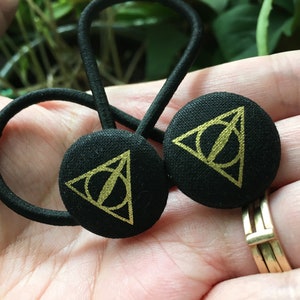 May include: Two black hair ties with circular fabric accents. Each accent features a gold-colored Deathly Hallows symbol. The hair ties are made of black elastic and are designed for securing hair. The background is blurred.