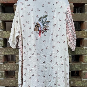 Könnte beinhalten: Cremefarbener Kimono-Cover-up mit floralem Muster und einem dekorativen Totenkopf- und Kopfschmuck-Patch auf dem Rücken. Die Ärmel haben ein gemustertes Design in Rosa- und Brauntönen. Der Cover-up hängt an einem schwarzen Kleiderbügel.
