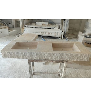 Peut inclure: Un grand évier double en pierre beige avec deux vasques rectangulaires. L'évier a un bord rugueux et texturé et est soutenu par un cadre métallique. Une machine étiquetée "MACH SPEED CNC S-7000" est en arrière-plan.