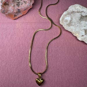 May include: A gold-coloured necklace featuring a heart-shaped pendant. The chain has a snake-like design. The necklace is displayed on a pink surface, with two decorative geodes in the background.
