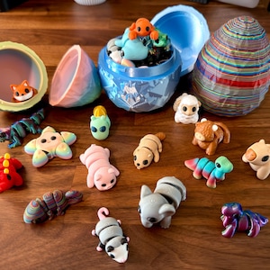 May include: A collection of colorful, small animal figurines, including a fox, sheep, and various other creatures. Some are inside decorative containers, including a blue egg-shaped container and a round container with a fox inside. The figurines are on a wooden surface.