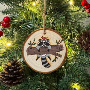 Op de afbeelding: Een houten kerstversiering met een handgeschilderde wasbeer die op een tak zit met een rode kardinaal erboven. De versiering is een natuurlijke houtschijf met een rustieke afwerking.