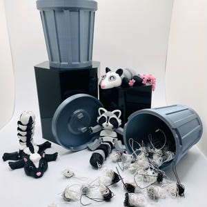 Op de afbeelding: Een grijze plastic prullenbak met een deksel, een wasbeerfiguur, een opossumfiguur en een stinkdierfiguur. De prullenbak is gevuld met witte en zwarte draad.