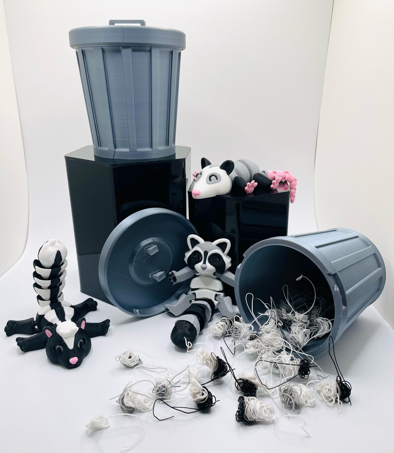Op de afbeelding: Een grijze plastic prullenbak met een deksel, een wasbeerfiguur, een opossumfiguur en een stinkdierfiguur. De prullenbak is gevuld met witte en zwarte draad.